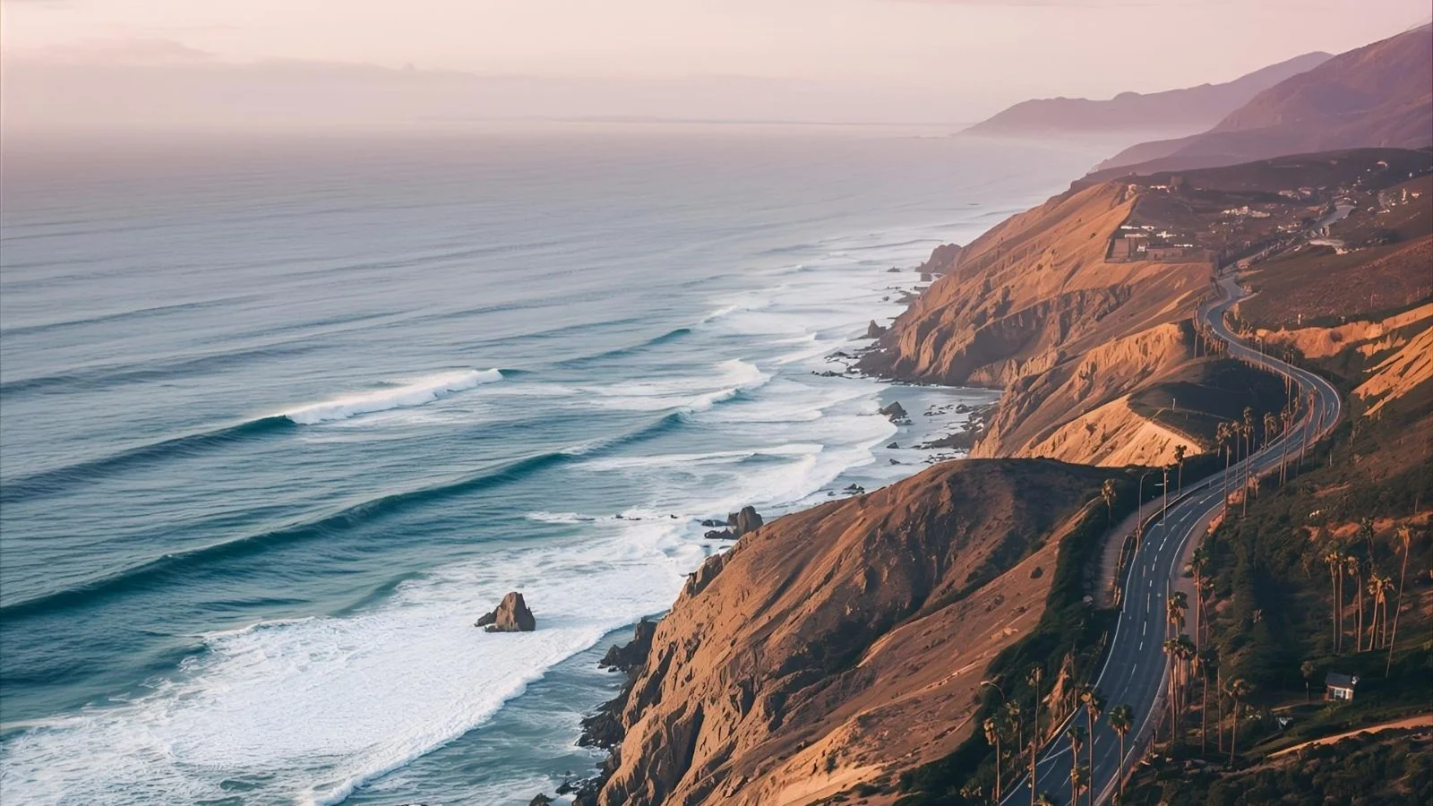 Golden hour aerial view of the California coastline with highway, cliffs, and ocean waves.